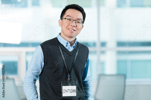 Smiling young Asian intern with a lanyard in an office, ideal for internship programs, entry level employment, or onboarding concepts.