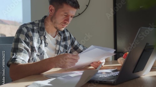 Worried man checking bills and financial papers while sitting at table with laptop. Stressed person planning home budget. Concept of personal finance review, expense control and household budgeting