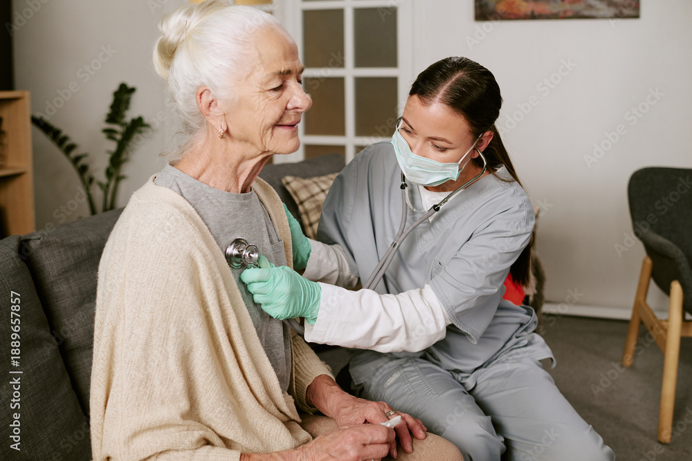 Obraz premium Senior Caucasian woman sitting on sofa receiving medical examination from young adult Asian female nurse wearing scrubs and mask using stethoscope in home setting