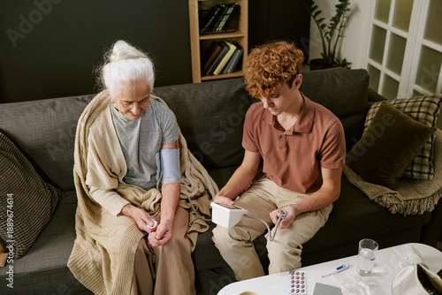 Senior Caucasian woman sitting on sofa having blood pressure measured by teenage Caucasian boy using digital monitor, both focused on health check, medication and water on table