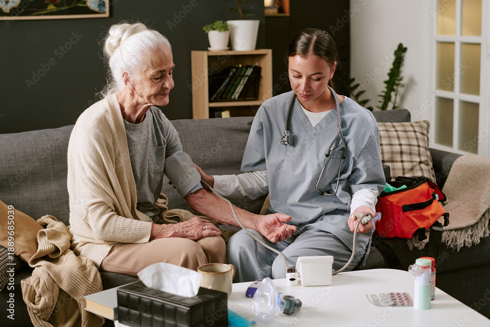 Obraz premium Senior Caucasian woman sitting on sofa receiving blood pressure measurement from young adult Caucasian female nurse using medical device, healthcare equipment visible on table