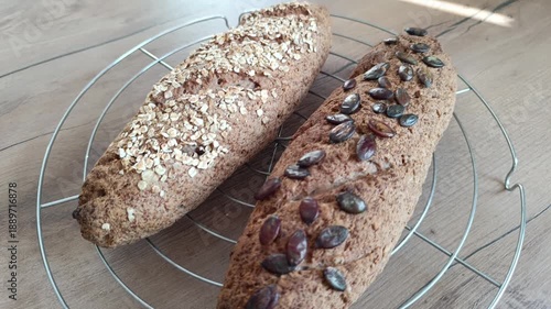 Horizontal close-up of two fresh homemade whole grain baguettes on a cooling rack. One loaf is topped with rolled oats and the other with pumpkin seeds. Healthy artisan bread on a wooden background.