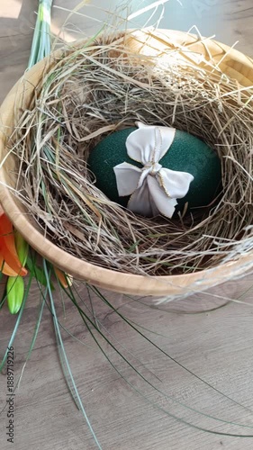 A vertical close-up of a large, emerald green decorative Easter egg with a white bow, nestled in a wooden bowl filled with dry straw. Vibrant orange lilies and green stems on a wooden surface.