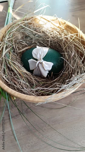 A top-down vertical shot of a shimmering emerald green Easter Emu egg decorated with a white satin bow, nestled in dry grass inside a wooden bowl. Elegant holiday composition with rustic texture.