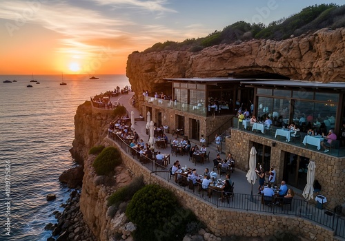 People enjoy a meal at a cliffside restaurant during sunset over the ocean