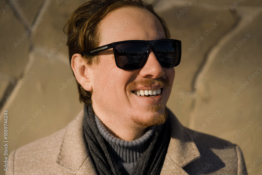 Obraz premium Close-up portrait of a young caucasian redhead man with sunglasses smiling wearing a coat on a winter day while walking down the street. Lifestyle.