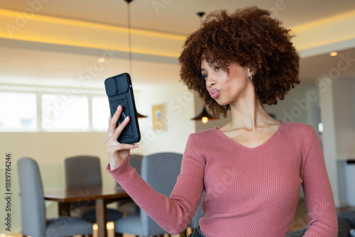 Female holding smartphone and posing with puckered lips in dining area wearing rose V-neck top