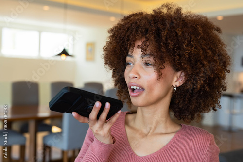 Woman in rose sweater speaking into smartphone held horizontally in dining area with wooden table