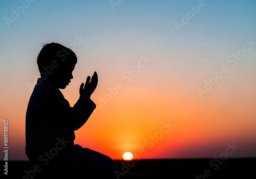 A devout young boy in silhouette raises his hands in prayer (Dua) against the dramatic, colorful sunset horizon.