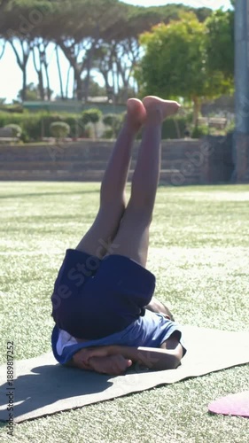 Vertical video: Cueing child in blue shirt and shorts barefoot doing core leg stretches on turf mat