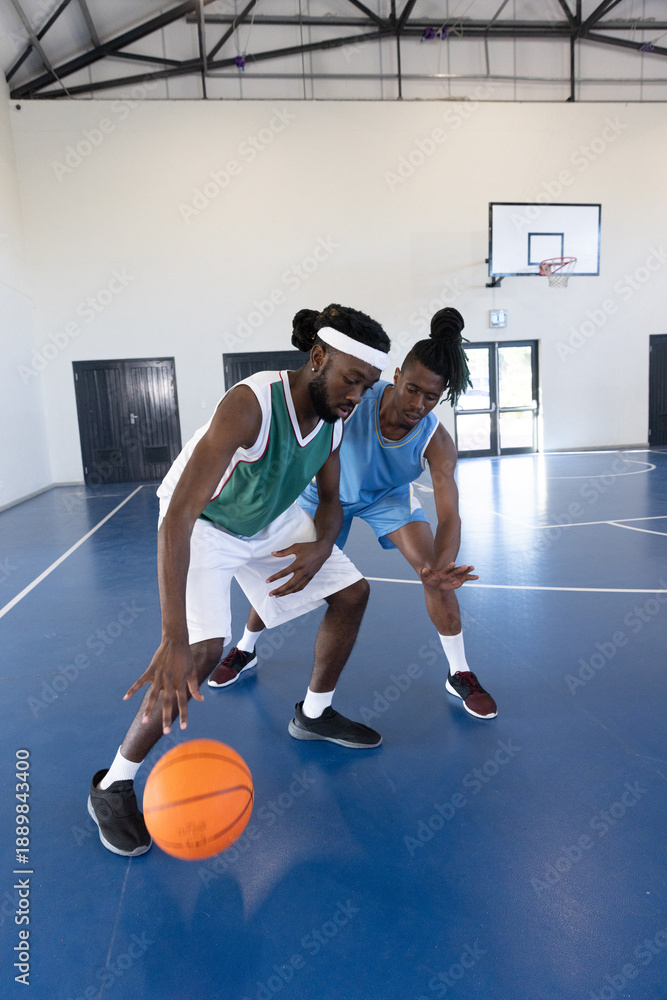 Fototapeta premium African american males dribbling and guarding basketball on court in gym near hoop by door