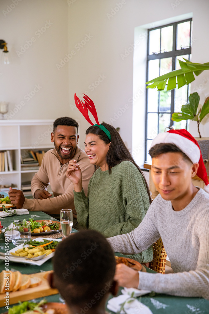 Naklejka premium Diverse friends laughing and chatting in home dining room with wine glasses, holiday headwear