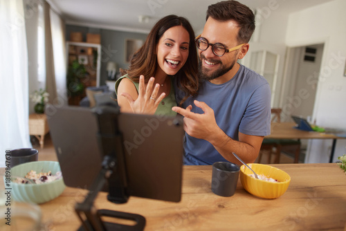 A happy couple joyfully shows off an engagement ring during a video call while enjoying breakfast together at their kitchen table, creating a warm and memorable moment.