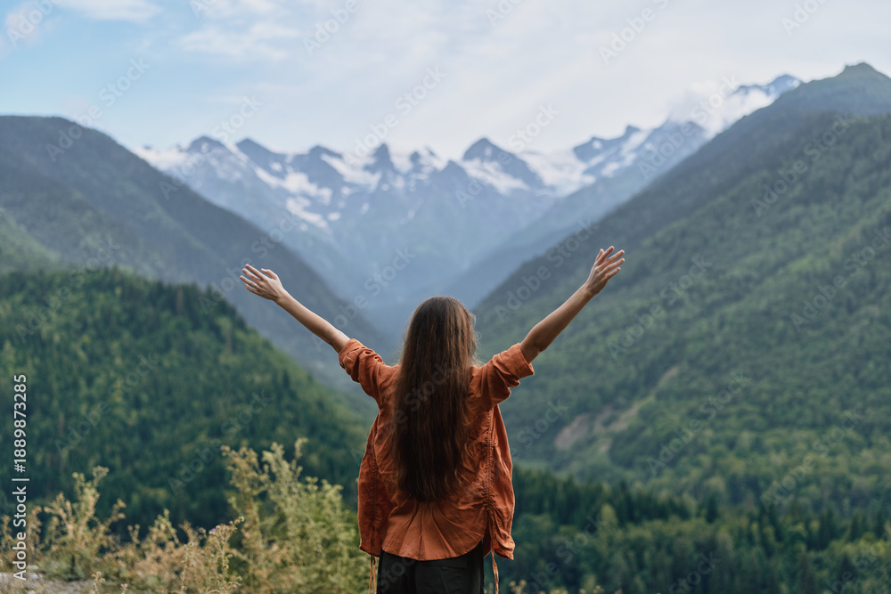 Obraz premium Woman with arms raised in a vast mountain landscape, embracing nature and freedom, gazing over green valleys toward snowy peaks under a clear alpine sky