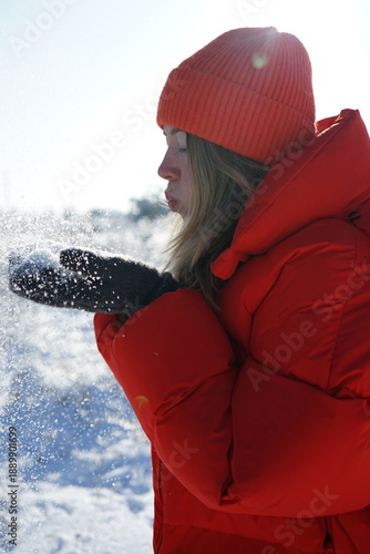 A girl blows snowflakes off her hands against the sun in a red jacket. 