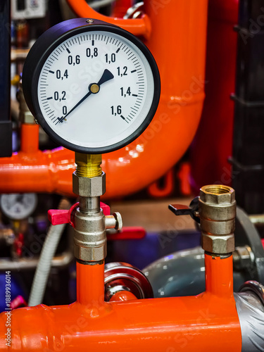 Pressure gauge on a bright orange pipe in an industrial setting with various equipment in the background during daytime