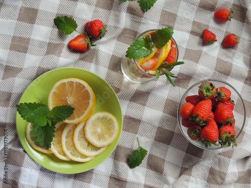 Ingredients for fruit lemonade on a checkered tablecloth and a glass with the drink, a flat lay, a homemade refreshing non-alcoholic cocktail with lemon, strawberries, mint leaves and lavender
