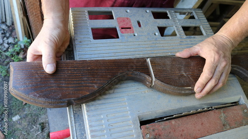Hands of a carpenter holding a prefabricated wooden piece with curved lines and varnished, cut into two parts, top view, showing the sawn wooden turned part of the furniture on the iron work surface