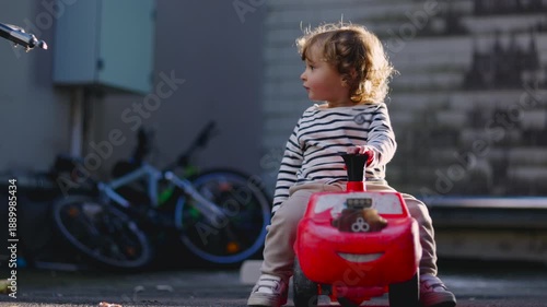 Toddler sitting on red toy car in corner of backyard courtyard turning slightly to side with focused expression creating quiet moment of attention curiosity and early awareness