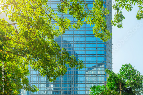 Fotografia Glass office building facade reflecting nearby towers framed by green trees Hong Kong city, China
