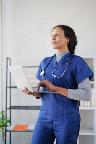 Female medical professional standing in an office, wearing blue scrubs and a stethoscope, looking away while typing on a laptop, representing healthcare innovation and technology