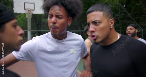 Hispanic men arguing on outdoor basketball court as African descent teammate intervenes to calm conflict, heated confrontation and emotions during street game