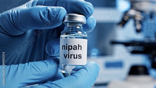 Close-up of a scientist in blue gloves holding a Nipah virus vaccine vial in a laboratory setting with a microscope. Concept of infectious disease research, medical breakthrough, and public health.