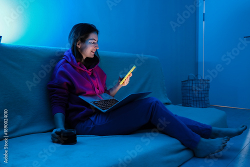 Young woman using smartphone and laptop in blue neon room light