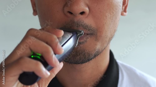 A man is shaving his mustache and beard with a razor in a closed room.