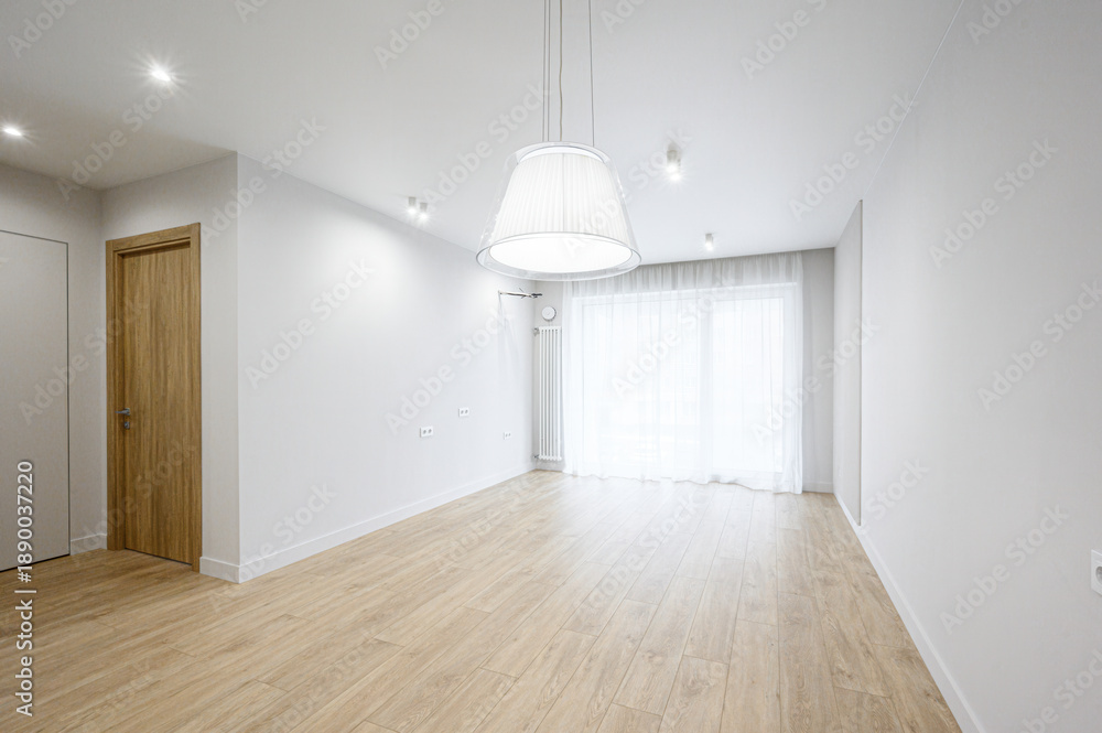 Obraz premium A bright, empty room with light wooden flooring, white walls, recessed ceiling lights, a large pendant lamp, sheer white curtains, and a wooden door — minimalist, airy space.