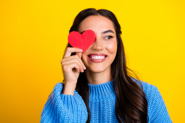 Joyful brunette woman in blue sweater smiles while holding a red heart over her eye against a yellow background creating a bright casual fashion lifestyle image