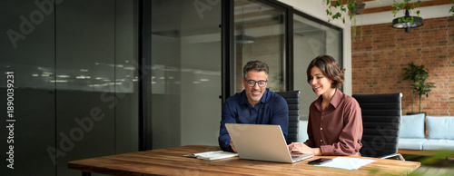 Female manager showing financial results to colleague at work. Two professional team business man and woman using laptop at corporate meeting. Happy busy executive people working together in office.
