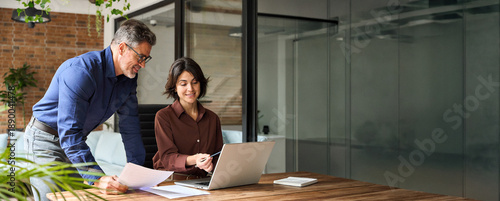 Two happy busy business leaders man and woman talking in office using laptop. Professional executive team people partners checking financial report working together on computer at corporate meeting.