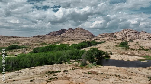 A panoramic view of rugged rocky mountains, a green valley with a small lake, and dense clouds creating a striking play of light and shadow.