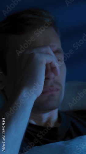 Young tired man with a phone in his hands scrolls through news feed or social media at night in his bedroom, but is interrupted by bad news or fatigue, disappointed and exhausted. Vertical video