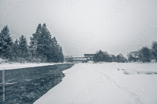Konstfotografi Bleak winter landscape with gray skies, snow-covered trees, partially frozen river, and a solitary bridge; cold, raw, moody, and desolate atmosphere