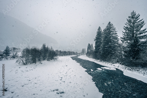 Photos Bleak winter landscape with gray skies, snow-covered trees, partially frozen river, and eerie silence; cold, raw, moody, and desolate atmosphere