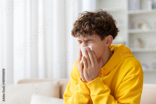 Valokuva Teenage boy sneezing into tissue at home, wearing yellow hoodie, showing cold or
