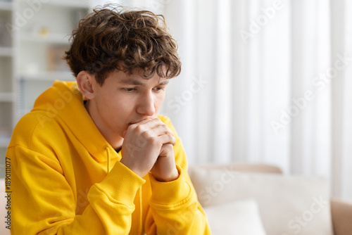 Foto Thoughtful teenage boy sitting on sofa with hands near face, wearing yellow hood