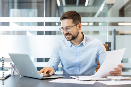 Young businessman engaged in professional work, holding paper document and typing on a laptop with a focused yet smiling expression in a bright, contemporary office environment