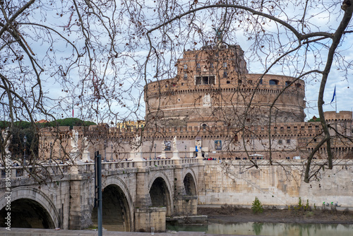 Wallpaper Mural Bare winter branches frame Castel Sant’Angelo and Ponte Sant’Angelo as visitors gather along the Tiber on a calm afternoon in Rome, Italy Torontodigital.ca