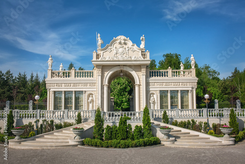 Elegant classical pavilion with sculpted statues and ornate archway stands within a formal garden, symmetrical steps and balustrades.