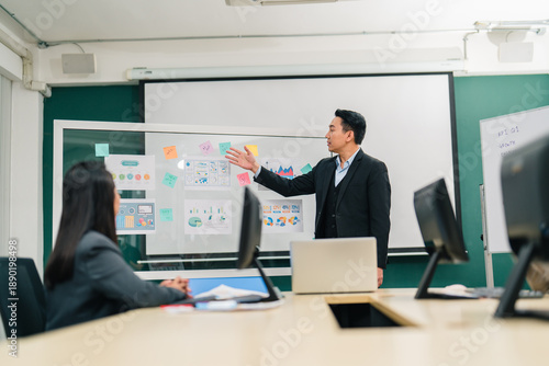 Serious Asian businessman points to financial charts and data on a visual board, presenting strategy and analysis to colleagues in a modern boardroom, leadership and planning