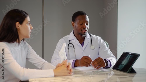 African American male orthopedic doctor discussing knee injury diagnosis with female patient using digital X-ray tablet and anatomical joint model in a modern medical clinic office.