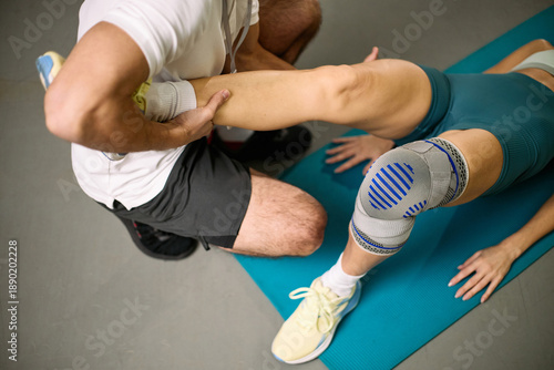 Caucasian young adult woman receiving physical therapy from Caucasian man therapist, lying on exercise mat while therapist supporting leg and knee during rehabilitation session
