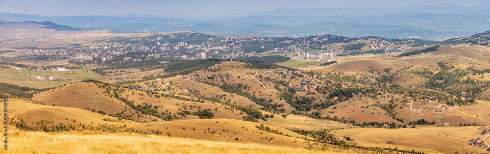 Fototapeta premium Panoramic view of Zlatibor landscape, featuring golden rolling hills, sparse vegetation, and a distant town under a pale sky