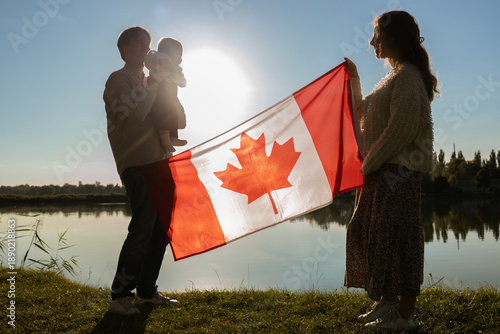 family of three with a large Canadian flag in the background at sunset. A man, a woman, and a small child in nature. A family outing on a holiday weekend. Happy Canada Day. Freedom, trust, pride