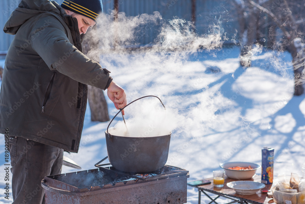 © T.Den_Team - Person preparing hot food outdoors during winter using a metal cauldron over open fire. Close view of hands cooking a meal in cold weather