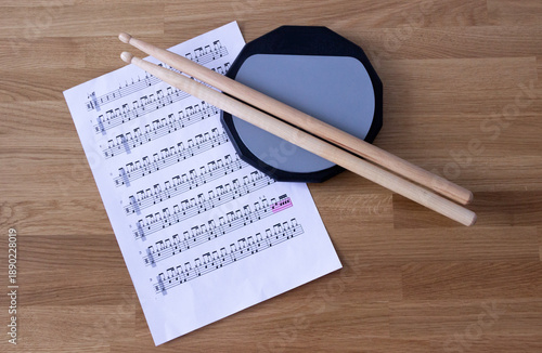 a pair of drums sticks on a grey practice pad and a music sheet on a wooden table, drums, drums practice, music education