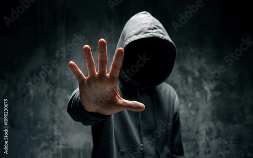Anonymous hooded man showing stop hand gesture in dark dramatic shadow